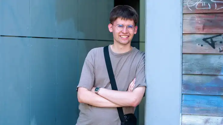 Ein junger Mann im T-Shirt steht mit verschränkten Armen angelehnt an eine Wand, die mit Graffiti besprüht.