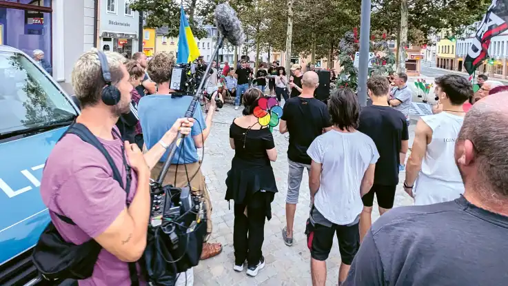 Auf einem kleinstädtischen Platz stehen ca. 40 Menschen im Kreis, in dessen Mitte ein junger Mann in ein Mikro spricht, im Vordergrund ein Kamerateam.