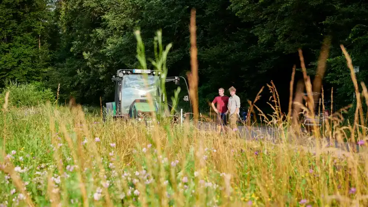  Zwei Männer stehen etwas fernab neben einem grünen Traktor und blicken auf eine satte Wildblumenwiese.