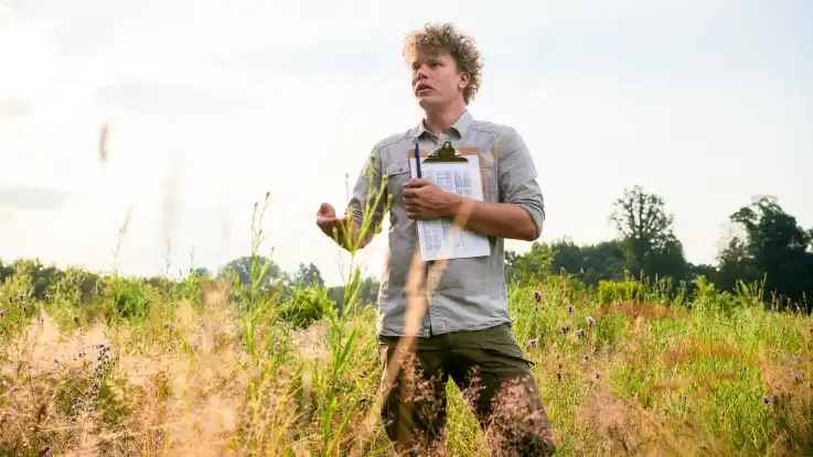 Ein junger Mann mit blonden Locken steht mit einem Klemmbrett auf einer dicht bewachsenen Wildblumenwiese.