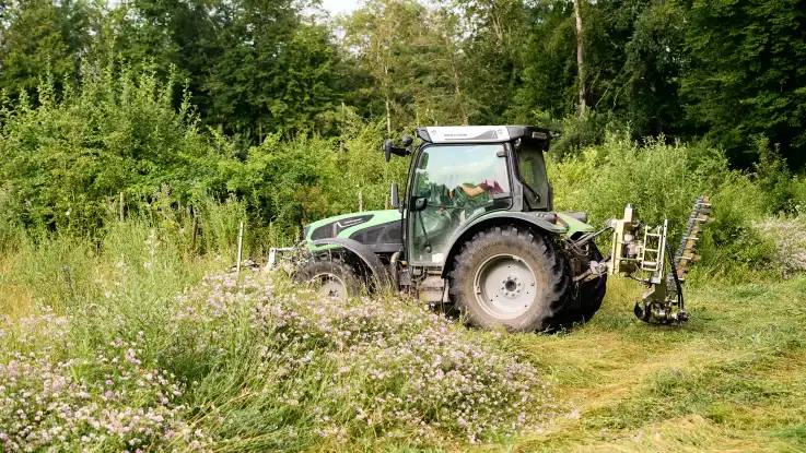 Der grüne Traktor ist nun seitlich zu sehen, an seinem Heck ist eine Apparatur aus Metall montiert.