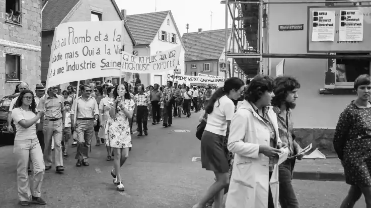 Ein bunt gemischter Protestzug mit französisch- und deutschsprachigen Plakaten bahnt sich seinen Weg durch eine Kleinstadt. 