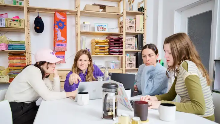 Die Frau im Overall sitzt mit drei weiteren Frauen an einem Tisch, auf dem Laptops und eine Kanne Kaffee stehen. 