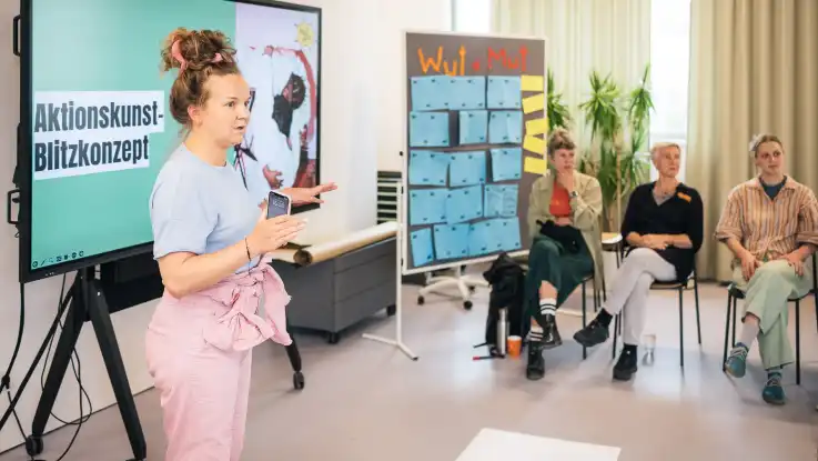 Eine Frau mit hochgesteckten Haaren und rosa Overall steht vor einem Smartboard und blickt fragend in die Runde. 