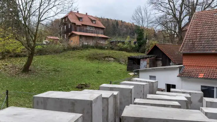 Neben einem Einfamilienhaus in ländlicher Kulisse stehen zahlreiche Quader in betongrau. 