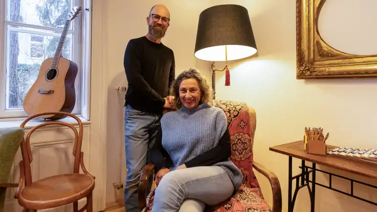 In einem Ohrensessel sitz eine junge Frau, hinter ihr anegelehnt an den Sessel steht ein junger Mann mit Vollbart. Beide lachen in die Kamera.