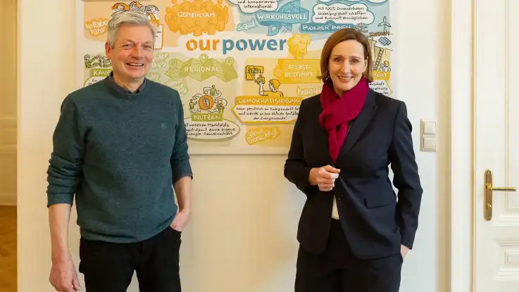 Vor einem OurPower Plakat an der Wand stehen die beiden Gründer: ein Mann mittleren Alters und eine jüngere Frau im Businessanzug.