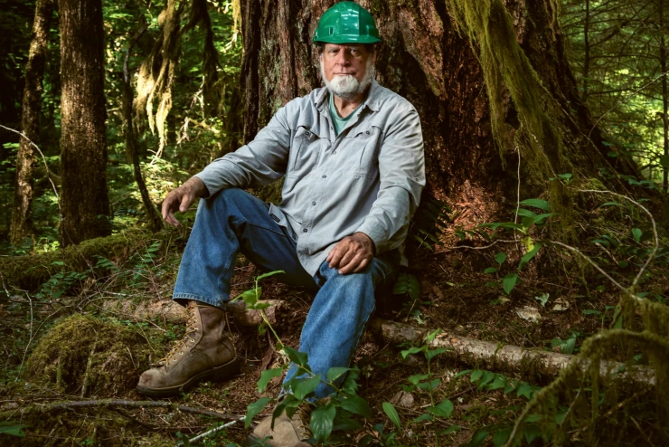 Ein Mann mit grauem Vollbart und grünem Schutzhelm sitzt in Arbeitskleidung an einem Baum gelehnt und schaut in die Kamera.