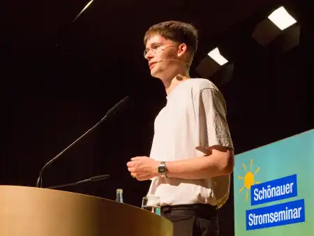 Ein junger Mann mit Brille und naturweißem T-Shirt steht an einem Rednerpult, hinter ihm eine Leuchtstele mit der Aufschrift «Schönauer Stromseminar»