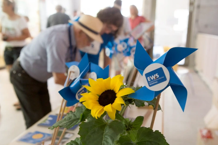 Eine Sonnenblume und kleine, blaue Spielzeugwindräder auf einem Tisch – dahinter sind unscharf Personen zu sehen. 