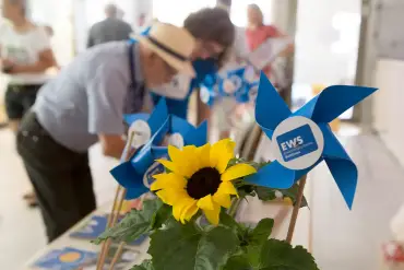 Eine Sonnenblume und kleine, blaue Spielzeugwindräder auf einem Tisch – dahinter sind unscharf Personen zu sehen. 