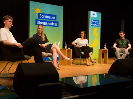 Vier Personen in lockerer Runde auf einem Podium, hinter ihnen Leuchtflächen mit den Aufschriften «Schönauer Stromseminar» und «Energie in Bürgerhand!»