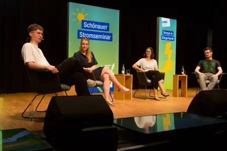 Vier Personen in lockerer Runde auf einem Podium, hinter ihnen Leuchtflächen mit den Aufschriften «Schönauer Stromseminar» und «Energie in Bürgerhand!»