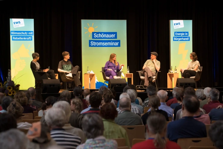 Vier Personen in lockerer Runde auf einem Podium, hinter ihnen Leuchtflächen mit den Aufschriften «EWS-Klimaschutz mit Rebellenkraft!», «Schönauer Stromseminar» und «EWS – Energie in Bürgerhand!»